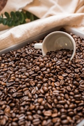 A white coffee cup is partially buried in a large pile of dark roasted coffee beans. The beans are glossy and densely packed, creating a rich texture. In the background, there are soft beige textiles and green leaves, adding a rustic and natural feel to the scene.