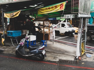A street food stall situated on a rainy urban sidewalk, with a woman in a raincoat standing next to it. The stall is covered with a green canopy and has a sign offering 'Sichuan cold noodles'. A blue cooler, scooter, and several small stools are nearby. In the background, vehicles including a white truck are visible on the street.