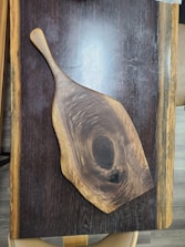A wooden cutting board with a natural, asymmetrical shape lies on a wooden table with visible grain patterns. The board displays rich textures and a polished surface, highlighting concentric wood rings in shades of brown. The table beneath it reflects a darker finish with contrasting lighter edges, suggesting a handcrafted aesthetic.
