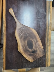 A wooden cutting board with a natural, asymmetrical shape lies on a wooden table with visible grain patterns. The board displays rich textures and a polished surface, highlighting concentric wood rings in shades of brown. The table beneath it reflects a darker finish with contrasting lighter edges, suggesting a handcrafted aesthetic.