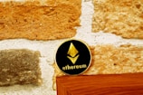 A coin with the Ethereum logo and text rests on a wooden surface against a textured stone wall. The coin has a black background with a gold Ethereum symbol and text.