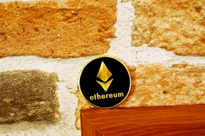 A coin with the Ethereum logo and text rests on a wooden surface against a textured stone wall. The coin has a black background with a gold Ethereum symbol and text.
