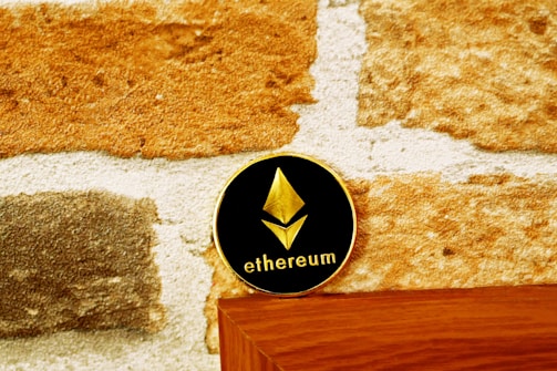 A coin with the Ethereum logo and text rests on a wooden surface against a textured stone wall. The coin has a black background with a gold Ethereum symbol and text.