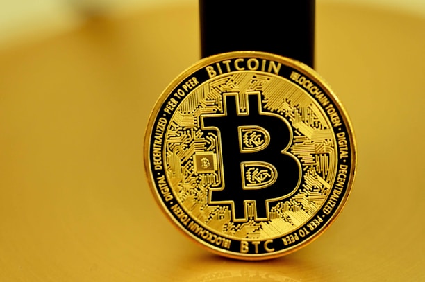 A golden coin featuring the Bitcoin symbol in the center, surrounded by intricate circuit-like designs. The coin bears the words 'Bitcoin' and 'Blockchain Token', along with other related terminology. The background is also golden, harmonizing with the color of the coin.