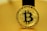 A golden coin featuring the Bitcoin symbol in the center, surrounded by intricate circuit-like designs. The coin bears the words 'Bitcoin' and 'Blockchain Token', along with other related terminology. The background is also golden, harmonizing with the color of the coin.
