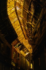 An industrial interior with a large, arched metal ceiling. The structure features intricate metal beams and girders, illuminated with warm lighting. The surrounding walls are dark, possibly made of wood or metal, giving a somewhat mysterious and abandoned feel.