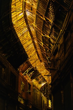 An industrial interior with a large, arched metal ceiling. The structure features intricate metal beams and girders, illuminated with warm lighting. The surrounding walls are dark, possibly made of wood or metal, giving a somewhat mysterious and abandoned feel.
