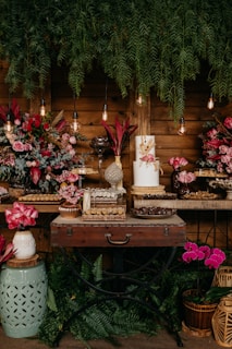 A rustic table setup with an arrangement of various colorful flowers, including pink and red roses, accompanied by a variety of decorative vases and glass containers. Hanging light bulbs provide a warm glow, while lush green foliage drapes from above. The table is adorned with a selection of sweets and a white cake with floral decorations.