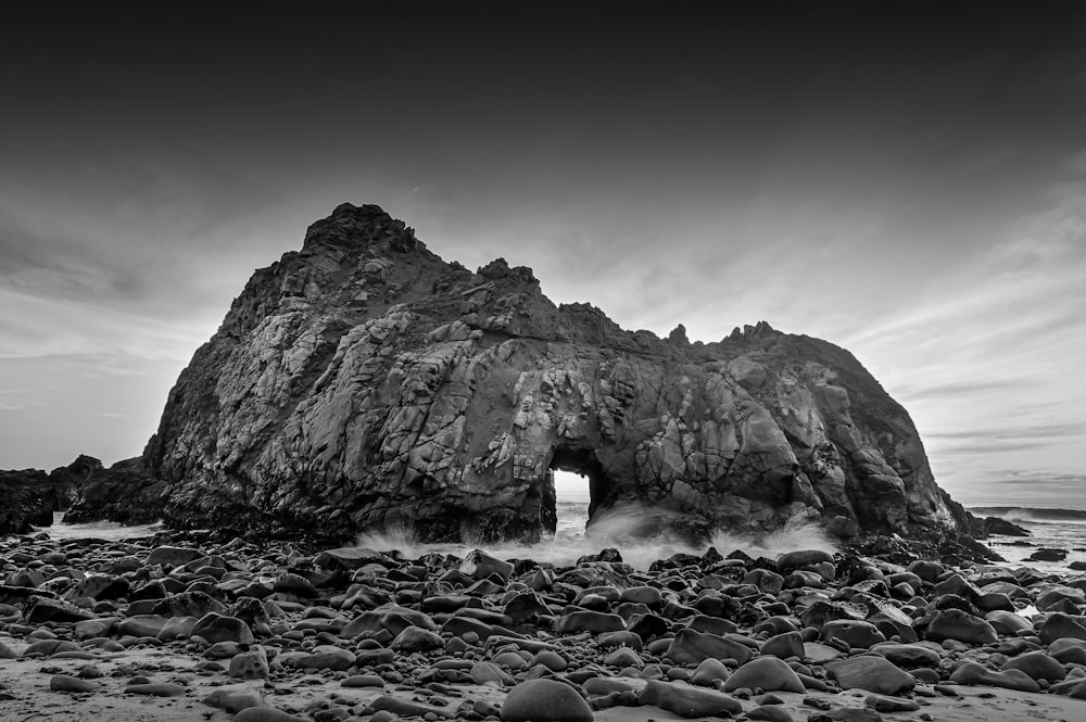 grayscale photo of person standing on rocky shore near rocky mountain