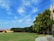 A spacious, well-maintained lawn is bordered by a row of trees with dense foliage. To the left, a yellow building with a red roof is visible. The sky overhead is a bright blue with scattered, wispy clouds. The foreground includes a paved pathway and landscaped greenery.