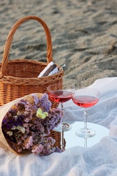 A wicker picnic basket sits on a sandy surface alongside a rolled towel. Nearby, two glasses filled with rosy-colored liquid rest on a reflective surface. A bouquet of pastel flowers, including purples and yellows, is placed on a light fabric.