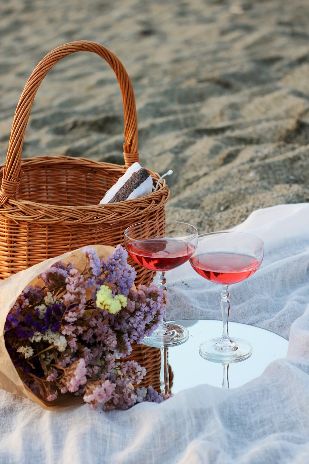 白いテーブルクロスに透明なワイングラスの横の茶色の編まれたバスケットにピンクと白の花