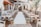 A beautifully arranged outdoor wedding venue with wooden chairs aligned on either side of an aisle covered in a white runner. The setting is embellished with elegant drapery and floral arrangements, creating an inviting and romantic atmosphere. Lanterns hang overhead, adding to the charm of the space. Greenery is strategically placed along the sides, and the backdrop features a decorative altar with more flowers.