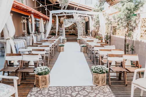 A beautifully arranged outdoor wedding venue with wooden chairs aligned on either side of an aisle covered in a white runner. The setting is embellished with elegant drapery and floral arrangements, creating an inviting and romantic atmosphere. Lanterns hang overhead, adding to the charm of the space. Greenery is strategically placed along the sides, and the backdrop features a decorative altar with more flowers.