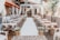 A beautifully arranged outdoor wedding venue with wooden chairs aligned on either side of an aisle covered in a white runner. The setting is embellished with elegant drapery and floral arrangements, creating an inviting and romantic atmosphere. Lanterns hang overhead, adding to the charm of the space. Greenery is strategically placed along the sides, and the backdrop features a decorative altar with more flowers.