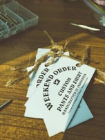 Several white tags tied with a brown twine are placed on top of a wooden surface. The tags read 'WEEKEND ORDER CUSTOM PANTS AND SHIRT HANDCRAFTED IN INDONESIA'. Nearby, there are small compartments holding metallic objects, possibly screws, and a pair of scissors.