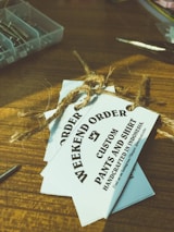 Several white tags tied with a brown twine are placed on top of a wooden surface. The tags read 'WEEKEND ORDER CUSTOM PANTS AND SHIRT HANDCRAFTED IN INDONESIA'. Nearby, there are small compartments holding metallic objects, possibly screws, and a pair of scissors.