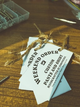 Several white tags tied with a brown twine are placed on top of a wooden surface. The tags read 'WEEKEND ORDER CUSTOM PANTS AND SHIRT HANDCRAFTED IN INDONESIA'. Nearby, there are small compartments holding metallic objects, possibly screws, and a pair of scissors.