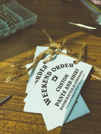 Several white tags tied with a brown twine are placed on top of a wooden surface. The tags read 'WEEKEND ORDER CUSTOM PANTS AND SHIRT HANDCRAFTED IN INDONESIA'. Nearby, there are small compartments holding metallic objects, possibly screws, and a pair of scissors.