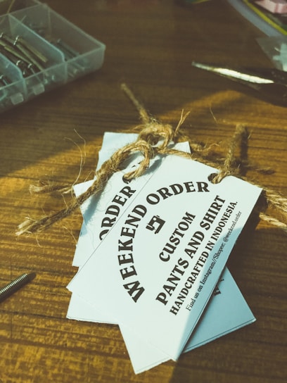 Several white tags tied with a brown twine are placed on top of a wooden surface. The tags read 'WEEKEND ORDER CUSTOM PANTS AND SHIRT HANDCRAFTED IN INDONESIA'. Nearby, there are small compartments holding metallic objects, possibly screws, and a pair of scissors.