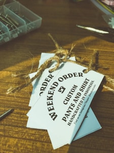 Several white tags tied with a brown twine are placed on top of a wooden surface. The tags read 'WEEKEND ORDER CUSTOM PANTS AND SHIRT HANDCRAFTED IN INDONESIA'. Nearby, there are small compartments holding metallic objects, possibly screws, and a pair of scissors.