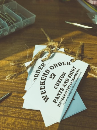 Several white tags tied with a brown twine are placed on top of a wooden surface. The tags read 'WEEKEND ORDER CUSTOM PANTS AND SHIRT HANDCRAFTED IN INDONESIA'. Nearby, there are small compartments holding metallic objects, possibly screws, and a pair of scissors.