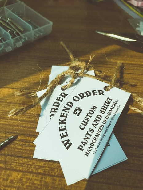 Several white tags tied with a brown twine are placed on top of a wooden surface. The tags read 'WEEKEND ORDER CUSTOM PANTS AND SHIRT HANDCRAFTED IN INDONESIA'. Nearby, there are small compartments holding metallic objects, possibly screws, and a pair of scissors.