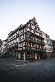 A traditional half-timbered building with intricate wooden framework stands prominently on a cobblestone street corner. The structure features large windows with decorative carvings and a steeply pitched roof. The surrounding area includes other historic buildings, contributing to a charming, old-world atmosphere.