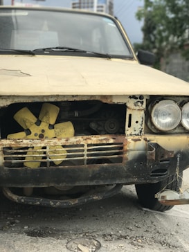 A vintage car with a heavily rusted front section, including its grille and bumper. The headlights are intact but aged, and a yellow fan is visible through the front due to the missing grille cover. The car is parked on a rough, concrete surface in an outdoor setting with some blurred buildings and greenery in the background.