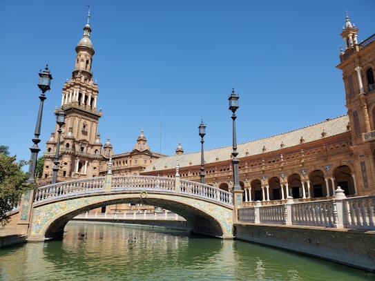 A stunning architectural site showcasing a grand building with a tall, ornate tower and intricate designs. A beautifully decorated bridge crosses over a green-colored waterway. The structure features multiple arches with detailed carvings, surrounded by elegant lampposts under a clear blue sky.