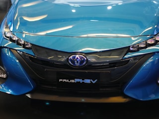 The image displays the front view of a blue Toyota Prius PHEV, highlighting its sleek headlights and prominent grille design.