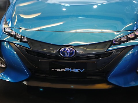 The image displays the front view of a blue Toyota Prius PHEV, highlighting its sleek headlights and prominent grille design.