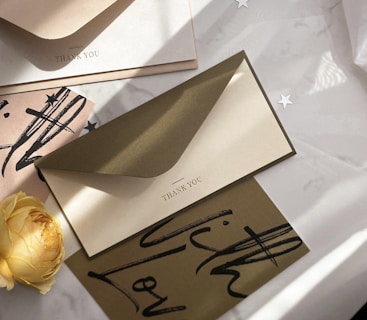 A collection of elegant thank you cards and envelopes arranged artfully on a surface. A single yellow rose adds a touch of softness to the scene. The cards and envelopes feature minimalistic text and artistic black brush strokes, creating a sophisticated and artistic atmosphere. Small star-shaped decorations are scattered around, contributing to a celebratory theme.