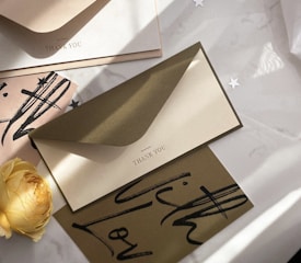 A collection of elegant thank you cards and envelopes arranged artfully on a surface. A single yellow rose adds a touch of softness to the scene. The cards and envelopes feature minimalistic text and artistic black brush strokes, creating a sophisticated and artistic atmosphere. Small star-shaped decorations are scattered around, contributing to a celebratory theme.