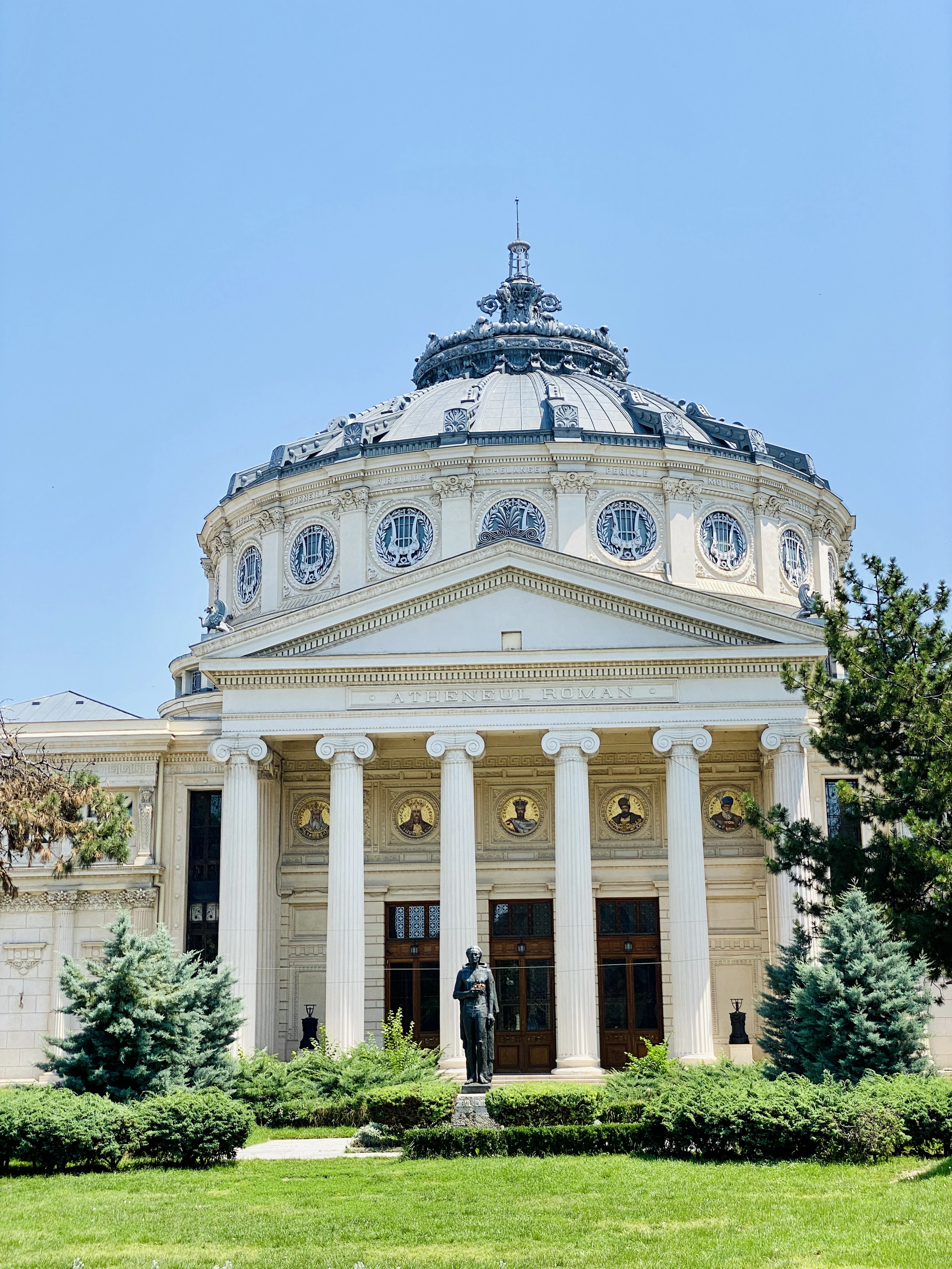 The Romanian Athenaeum in Bucharest under a clear blue sky, its Ionic portico inscribed Ateneul Roman below the ribbed dome and its ring of lyre medallions, with a bronze statue in front and clipped conifers on the lawn.
