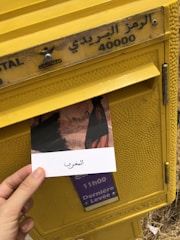 A hand holding a postcard depicting a scene, with text in Arabic, is being placed into a yellow postal box. The box has Arabic lettering and the number 40000 on it, along with a sign indicating the last collection time. The surroundings suggest an outdoor setting.