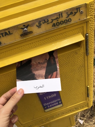 A hand holding a postcard depicting a scene, with text in Arabic, is being placed into a yellow postal box. The box has Arabic lettering and the number 40000 on it, along with a sign indicating the last collection time. The surroundings suggest an outdoor setting.