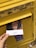 A hand holding a postcard depicting a scene, with text in Arabic, is being placed into a yellow postal box. The box has Arabic lettering and the number 40000 on it, along with a sign indicating the last collection time. The surroundings suggest an outdoor setting.