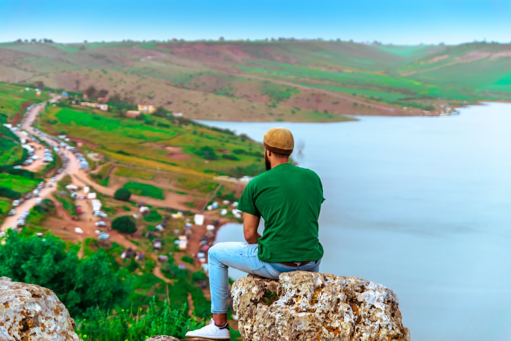 man in green shirt and blue denim jeans sitting on rock near body of water during