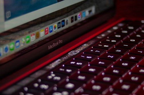 A close-up image of a MacBook Pro laptop focusing on the red backlit keyboard with illuminated keys. The screen displays a row of application icons on the dock, including well-known software. The angle highlights both the sleek design of the keyboard and a portion of the monitor.