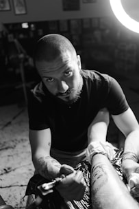 A dark-themed photo of a tattoo artist carefully working on a client's arm in a cozy studio.