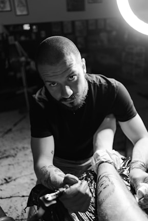 A tattoo artist works intently on a client's arm, creating intricate designs while surrounded by a dimly lit studio. The focus is on the artist's expression and the details on the client's tattoo.
