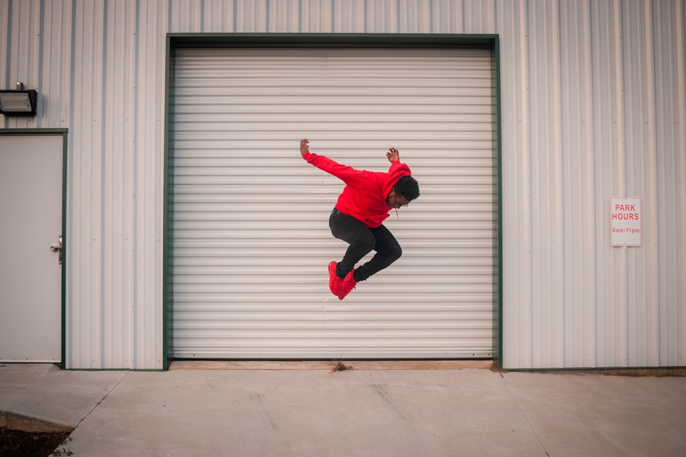 Hombre con chaqueta roja y pantalones negros saltando cerca de la puerta enrollable blanca
