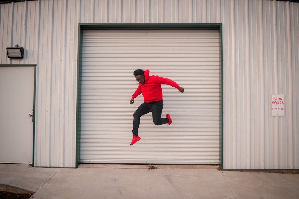 man in red long sleeve shirt and black pants jumping near gray roll up door during