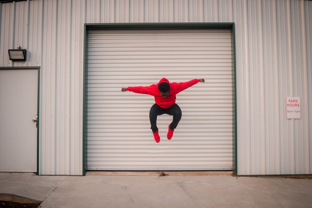 灰色のロールアップドアの前でジャンプする赤いジャケットの男
