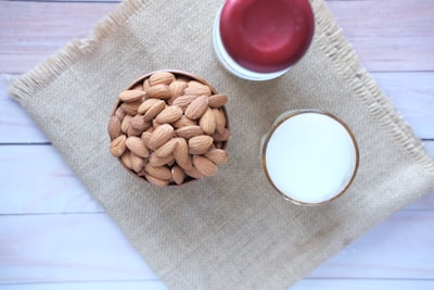 A bowl filled with whole almonds sits on a piece of burlap material placed on a light wooden surface. Next to the bowl is a glass of milk and a jar with a red lid.