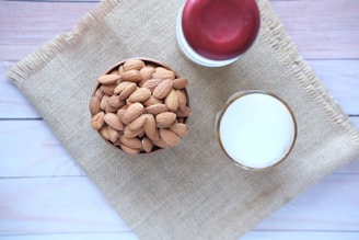A bowl filled with whole almonds sits on a piece of burlap material placed on a light wooden surface. Next to the bowl is a glass of milk and a jar with a red lid.