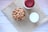 A bowl filled with whole almonds sits on a piece of burlap material placed on a light wooden surface. Next to the bowl is a glass of milk and a jar with a red lid.