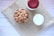 A bowl filled with whole almonds sits on a piece of burlap material placed on a light wooden surface. Next to the bowl is a glass of milk and a jar with a red lid.