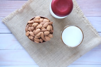 A bowl filled with whole almonds sits on a piece of burlap material placed on a light wooden surface. Next to the bowl is a glass of milk and a jar with a red lid.